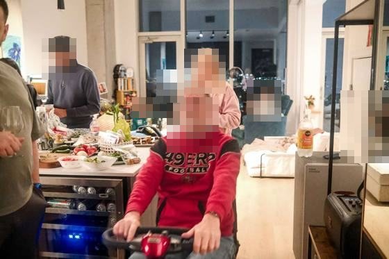 Man grinning while riding a toy scooter through a party kitchen, guests reacting behind