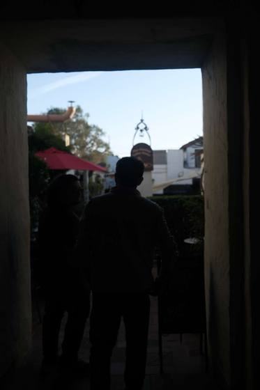 Two figures silhouetted in a dark adobe passage looking out to a sunlit street