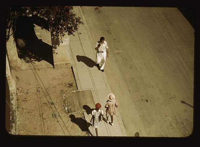 Overhead 1940s Kodachrome of a sunlit sidewalk: long shadows, a man in white walking toward two children